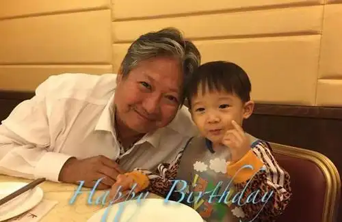 照片上,洪金宝穿着一件白色衬衫,抿嘴微笑,低着头和孙子紧挨在一起,而