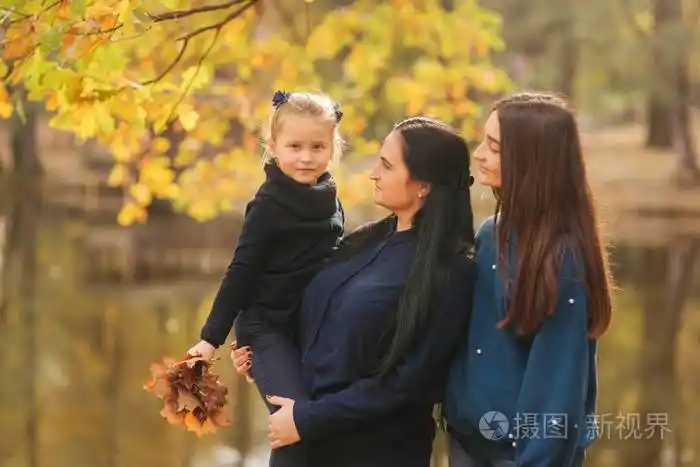 妈妈和两个女儿在公园玩得很开心最小的女儿收集树叶和微笑愉快的家庭