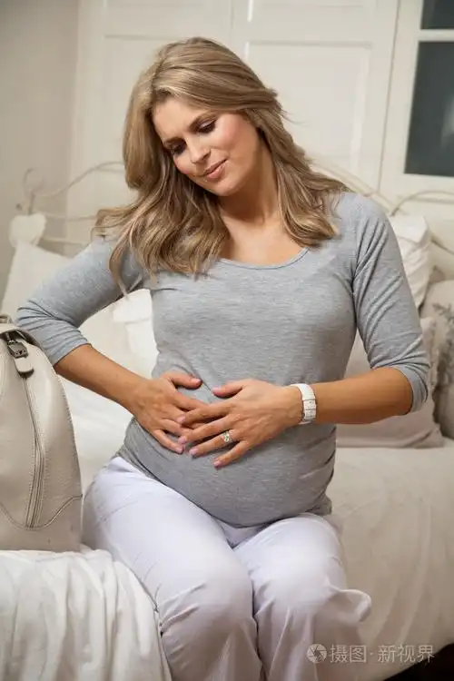 怀孕的妇女坐在床上与收缩疼痛和休息的双手放在她的肚子上照片-正版