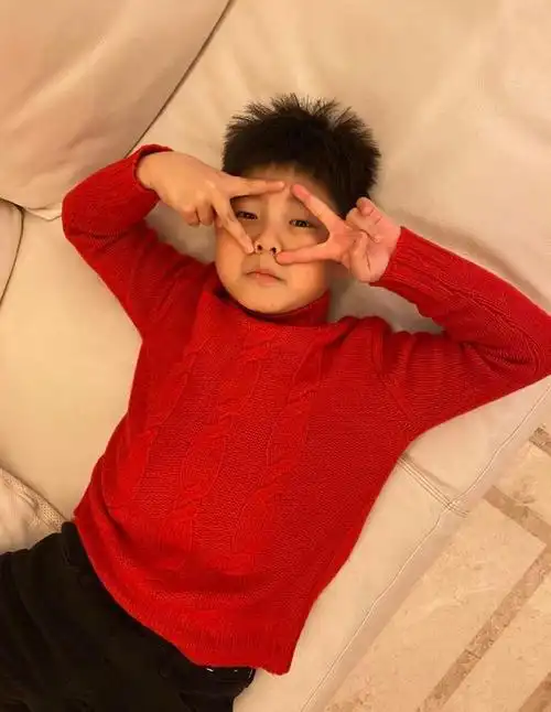 安吉穿着红色毛衣,理着一个刺猬头,躺在沙发上,双手在眼睛旁边比耶