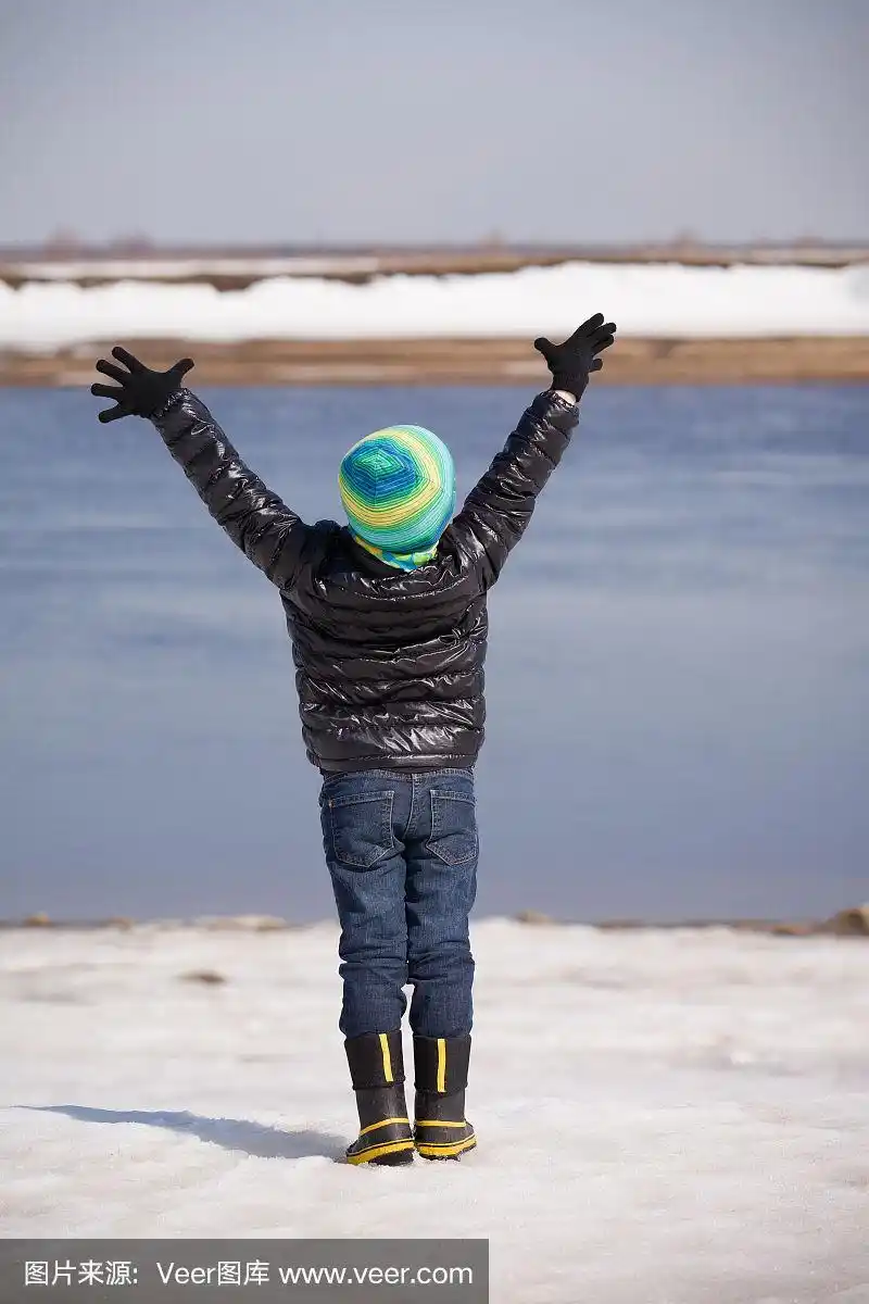 一个可爱的快乐的孩子男孩的背影站在河岸边与他的手在阳光明媚的冬天