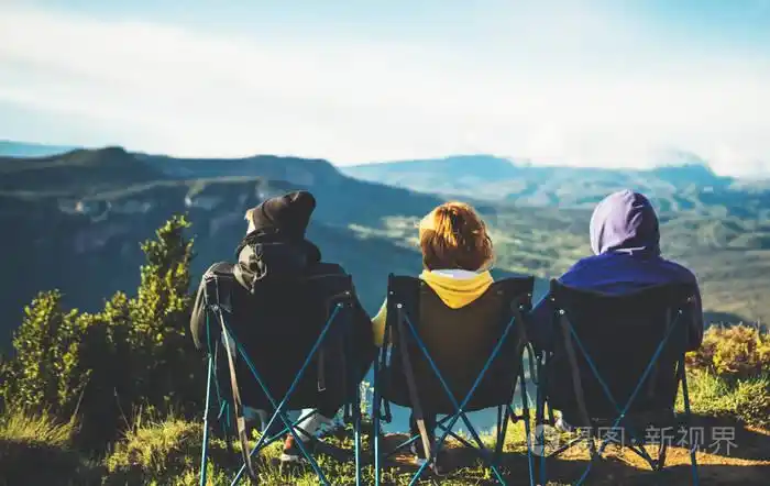 三个朋友坐在山顶的野营椅上享受大自然,拥抱游客,在全景景观周末概念