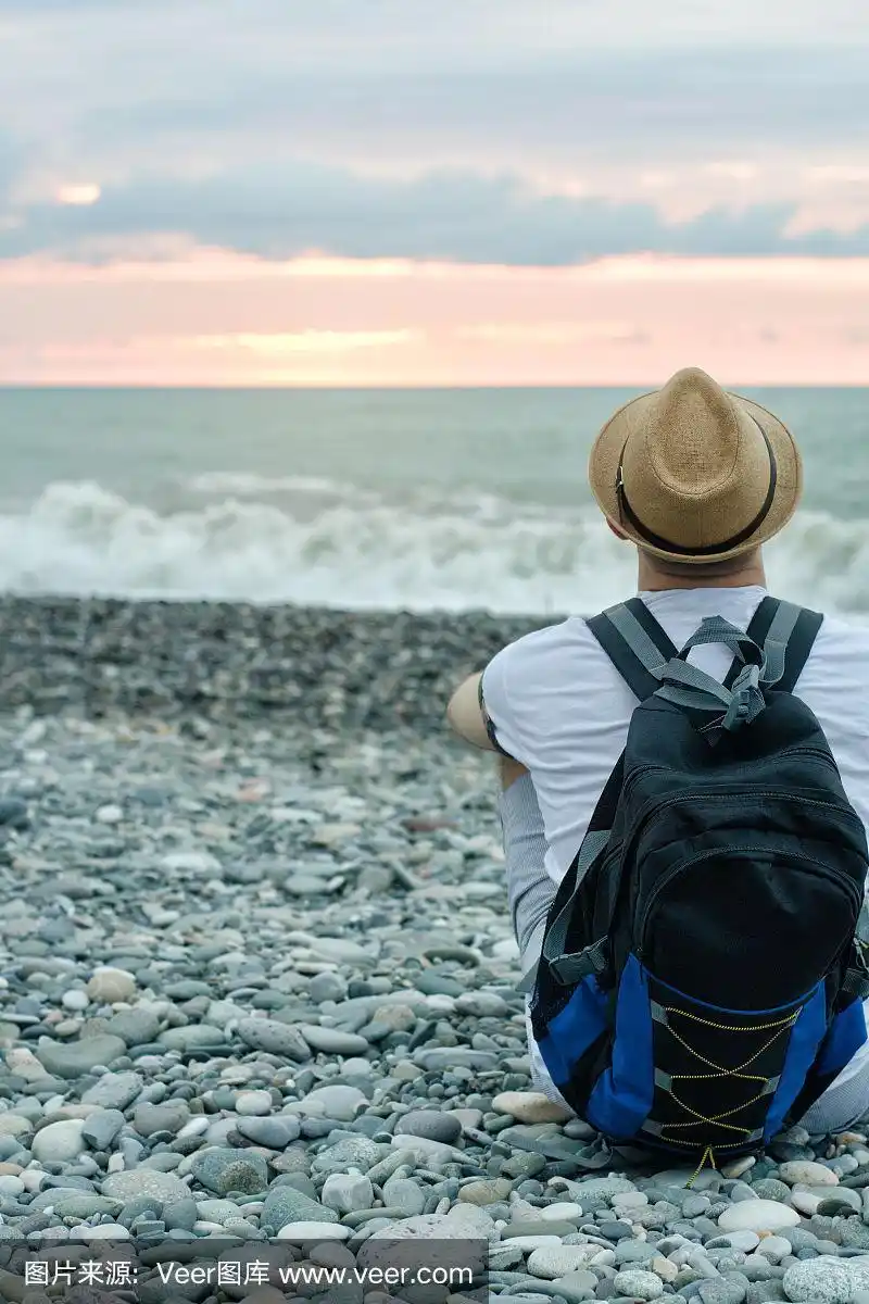 一个戴着帽子背着背包的人坐在海滩上,背对着大海和日落的天空.后视图