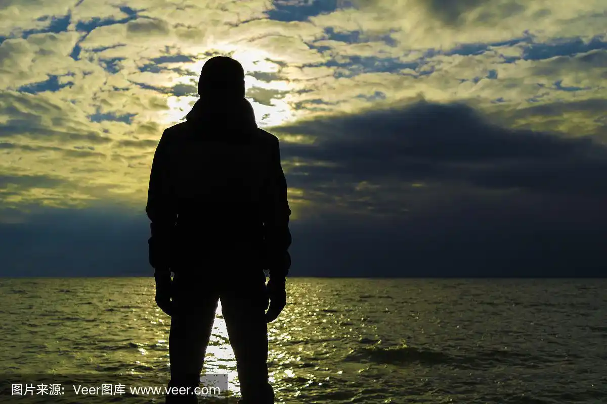 穿着夹克和帽子的孤独男子的剪影站在木制海岸上,看着海洋上的日落.