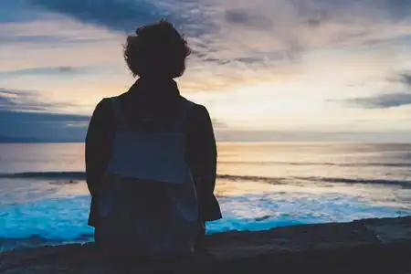 戴着帽子,望着大海的背影快乐的年轻人穿着t恤坐在海边,看着大海,背影