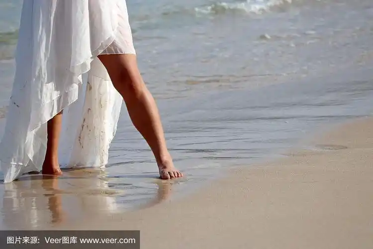光着脚的女孩穿着白色的裙子在沙滩上抹着沙子走在海浪的背景上