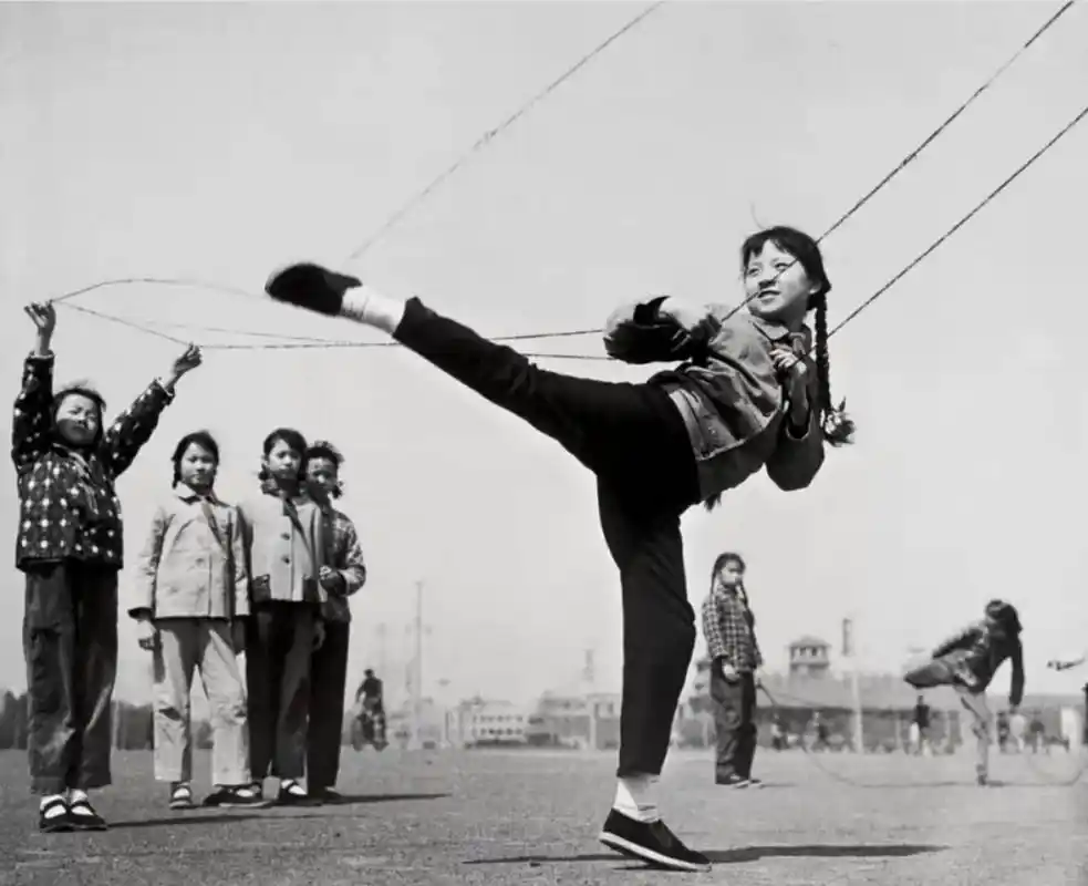 照片中,我们看到一位女同学在操场上跳皮筋,周围是那个时代的建筑物和