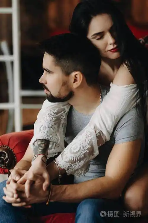 坐在一张红色的椅子上,有纹身和红唇的女孩正从背后拥抱着他的男人,他