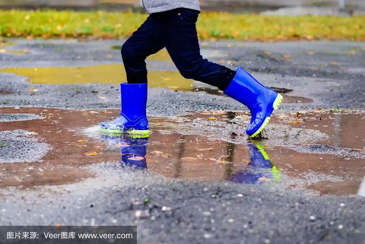 穿着蓝色橡胶雨靴的男孩跳到肮脏的水坑里