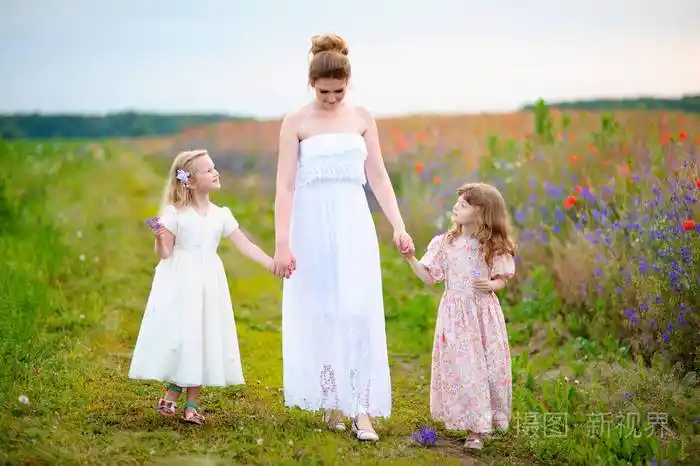 漂亮的母亲穿着白色的礼服与两个小女孩步行通过夏季场