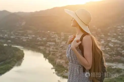 背上背包和帽子的漂亮女孩侧身站在城市和下面的河里的背景
