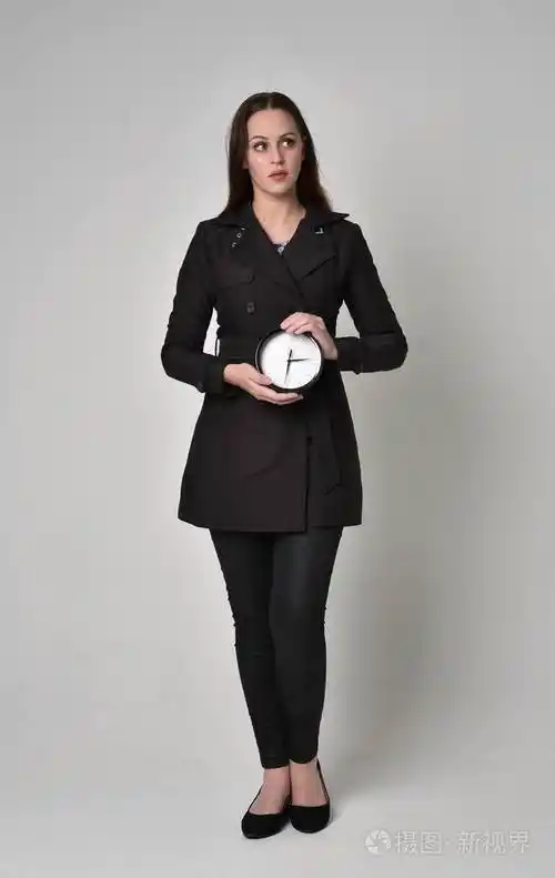 一个黑发女孩穿着黑色长外套站在灰色工作室背景上的姿势的全长肖像