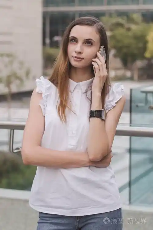 年轻的欧洲女性与棕色头发, 在白色上衣和蓝色牛仔裤打电话通过手机与