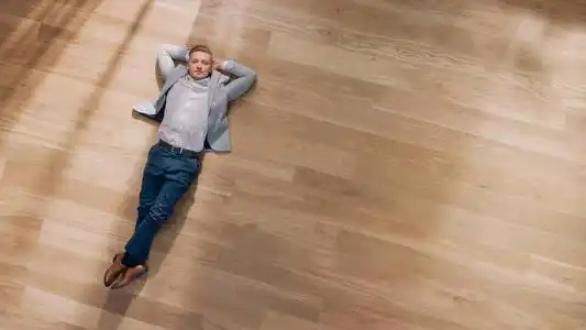 穿着棕色套头衫和灰色牛仔裤的年轻人躺在沙发上,用笔记本电脑.