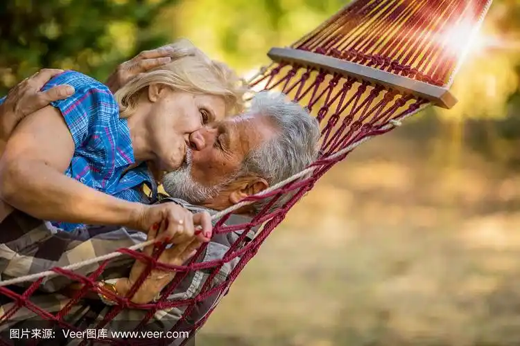 一对情侣在吊床上接吻.一对老年夫妇在红色条纹吊床上微笑放松.