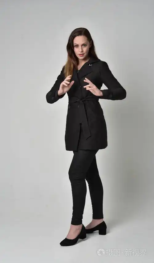 一个黑发女孩穿着黑色长外套站在灰色工作室背景上的姿势的全长肖像.