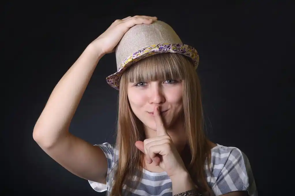 可爱的年轻微笑女孩用手势"嘘"和保持的帽子