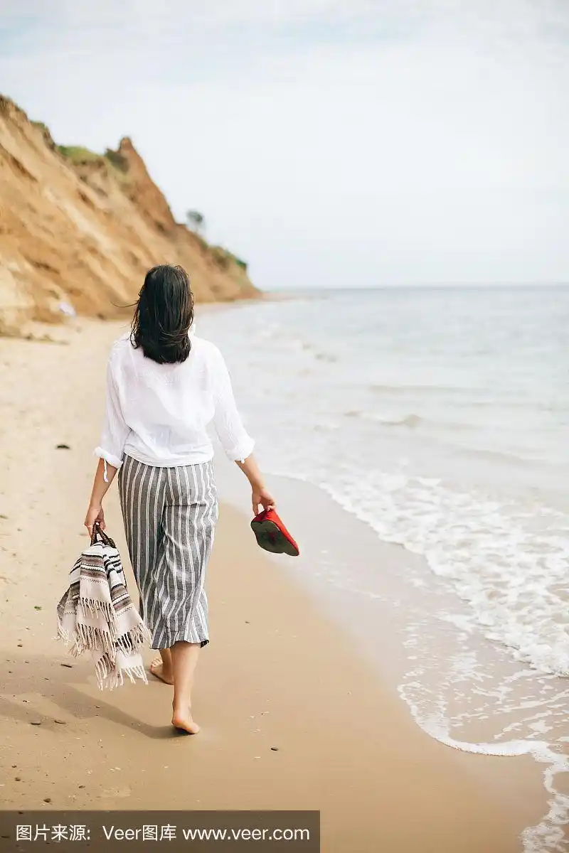 时髦的时髦女孩赤脚走在沙滩上,手里拿着包和鞋.