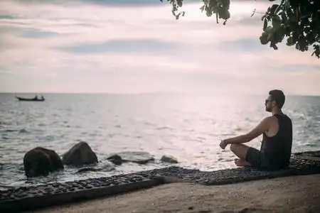 一个年轻人坐在海滩上. 戴太阳镜的帅哥在日落时看着海洋上的地平线.