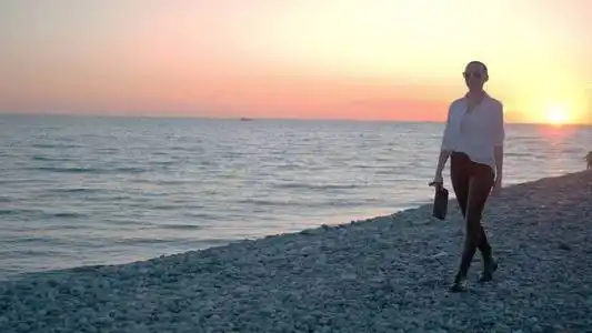 一个穿着白衬衫,皮裤和靴子的女人沿着海边散步,欣赏着日落.照片