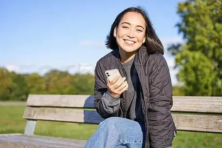 可爱的年轻女子手里拿着智能手机,坐在长凳上,微笑着,用手机,在公园里