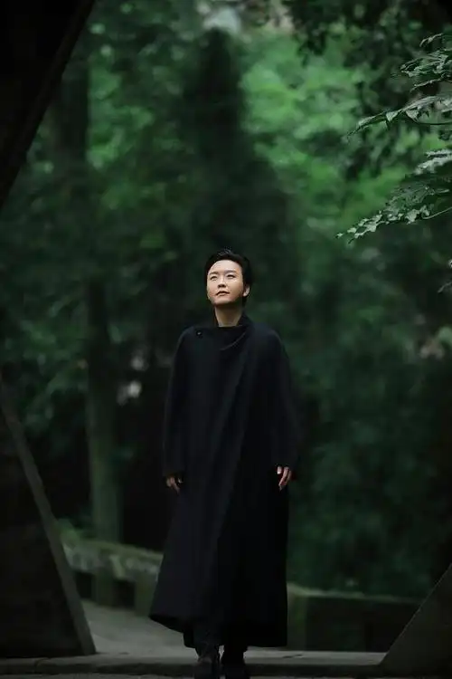 写真中李玉刚身着一身黑色长袍,简简单单的装扮,行走在石像林立,青葱