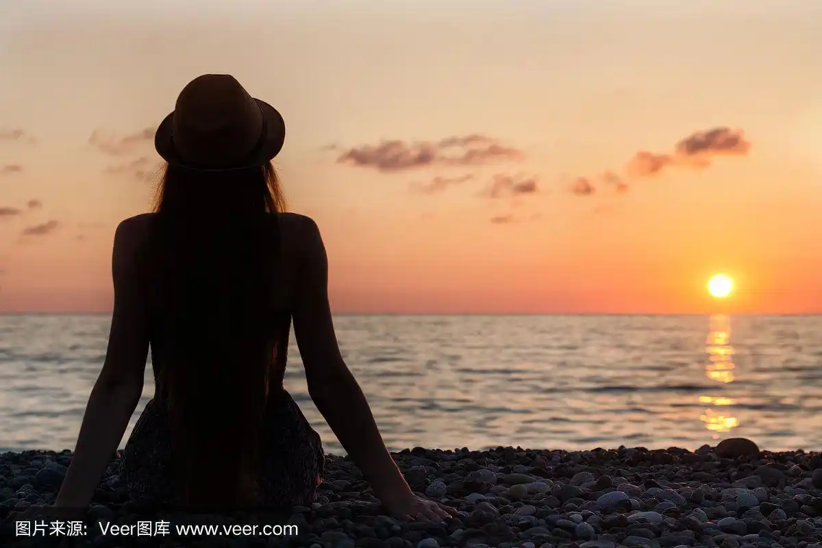 戴着帽子的女孩坐在海边.日落时间.轮廓.后视图