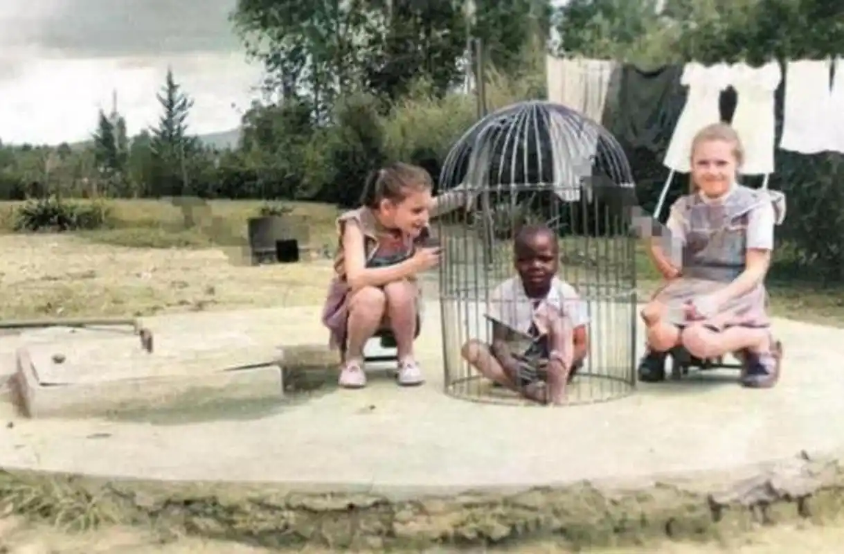 1958年比利时,两个白人小女孩围观着一个被关在笼子里的黑人小男孩笑