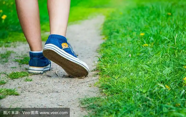 走路的女人腿在牛仔裤运动鞋的道路和绿色的草地上