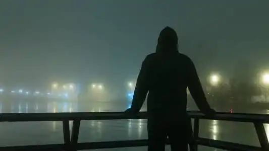 一个戴着兜帽的人在河边的路灯下剪影.在一个神秘的,雾蒙蒙的冬夜照片